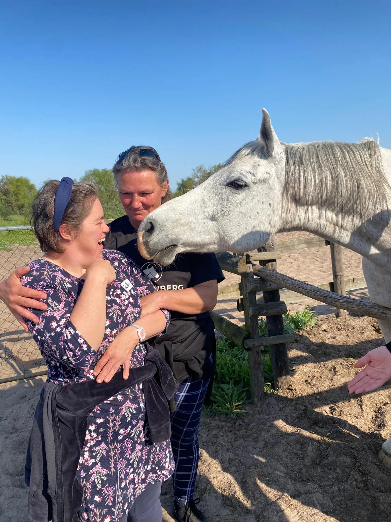A resident laughing with delight beside a grey horse leaning over the paddock rail, supported by a therapist