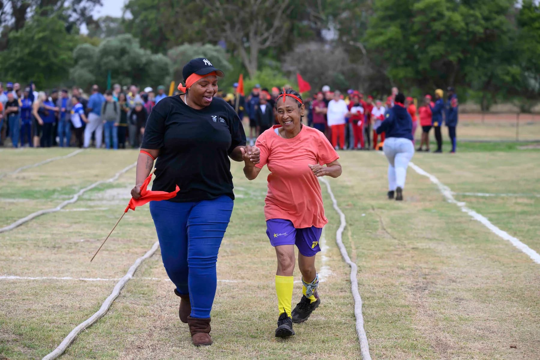 A resident and a staff member running hand in hand down the marked lanes of the sports field while the rest of the village cheers